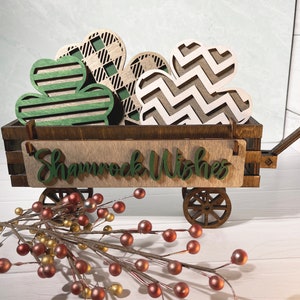 May include: A wooden wagon filled with heart-shaped decorations. The hearts have various patterns, including stripes, plaid, and zigzags. The wagon has a sign that reads "Shamrock Wishes" in green script. A sprig of berries is in the foreground.