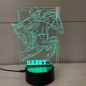 May include: A neon green LED night light depicting a horse and jockey. The name "GABBY" is lit up at the base. The light is set on a black circular base with a power cable.