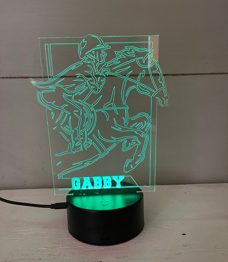 May include: A neon green LED night light depicting a horse and jockey. The name "GABBY" is lit up at the base. The light is set on a black circular base with a power cable.