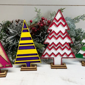 May include: Four decorative wooden Christmas tree ornaments on stands. The first is a natural wood tree with a magenta awareness ribbon. The second has yellow and purple horizontal stripes. The third is white with red zigzags. The fourth is green with white holly leaves and red berries.