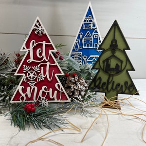 May include: Three decorative wooden Christmas tree ornaments. One is red with white text reading Let it snow and a snowflake. One is blue with a white village pattern. One is olive green with a black nativity scene and the word Believe.