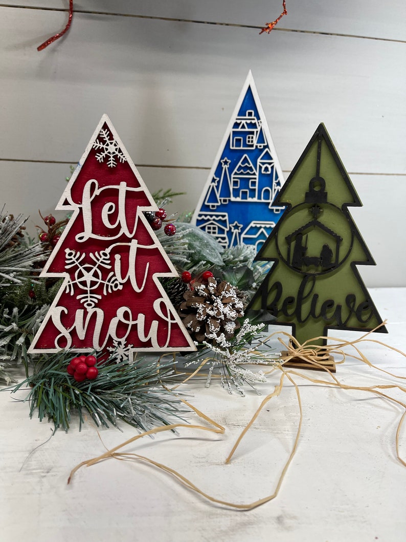 May include: Three decorative wooden Christmas tree ornaments. One is red with white text reading Let it snow and a snowflake. One is blue with a white village pattern. One is olive green with a black nativity scene and the word Believe.