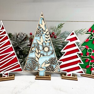May include: Four decorative wooden tree cutouts standing on small rectangular bases. The first is red with white diagonal lines, the second features a light blue background with intricate floral cutouts, the third has red and white stripes, and the fourth is green with red light bulb shapes.
