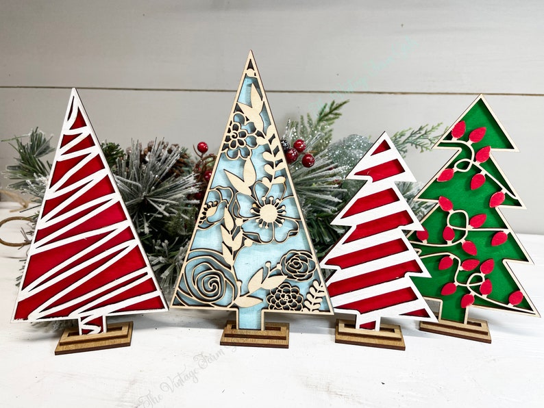 May include: Four decorative wooden tree cutouts standing on small rectangular bases. The first is red with white diagonal lines, the second features a light blue background with intricate floral cutouts, the third has red and white stripes, and the fourth is green with red light bulb shapes.