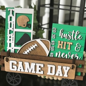 May include: A wooden wagon with football-themed decorations. The wagon features a football, helmet, and the words "Game Day." Additional signs read "Football" and "Hustle, Hit & Never Quit." The color scheme includes green, brown, and white.