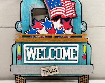 Op de afbeelding: Een houten decoratie in de vorm van een truck met een patriottisch thema. De truck is lichtblauw geschilderd met een grijze bumper en zwarte banden. Het heeft een Amerikaanse vlag, rode, witte en blauwe sterren en het woord "WELCOME".