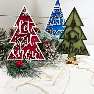 May include: Three decorative tree-shaped ornaments. The red one features the text Let it snow and snowflake cutouts. The blue one displays a white village scene. The olive green one shows a nativity silhouette and the word Believe.