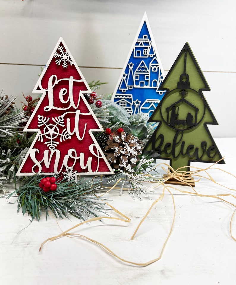 May include: Three decorative tree-shaped ornaments. The red one features the text Let it snow and snowflake cutouts. The blue one displays a white village scene. The olive green one shows a nativity silhouette and the word Believe.