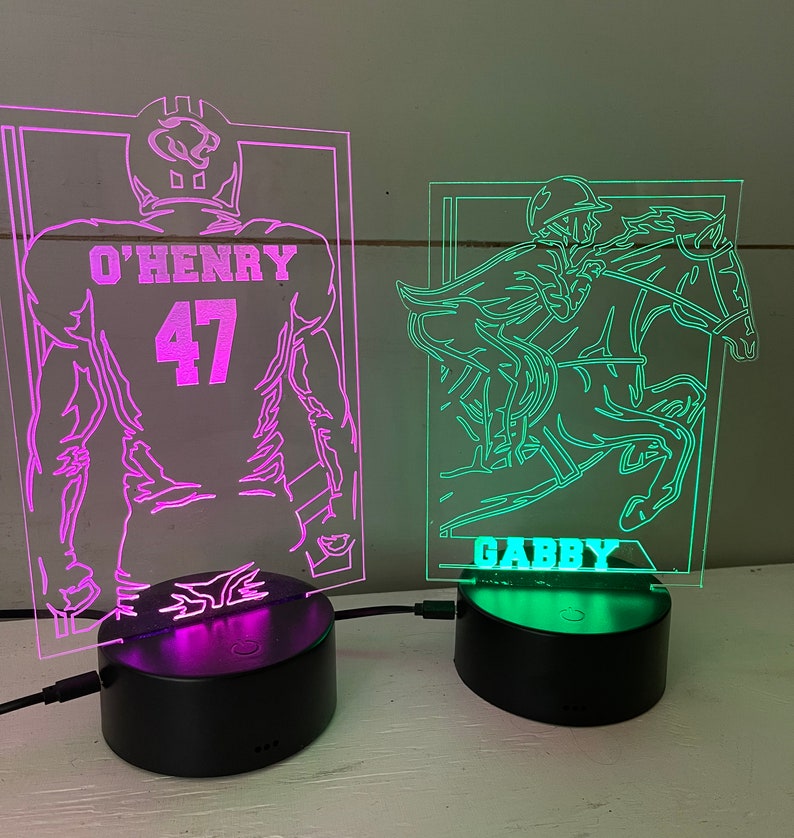 May include: Two illuminated acrylic lamps with black bases. One features a football player with the name "O'HENRY" and the number "47". The other depicts a jockey on a horse with the name "GABBY". Both lamps have a neon glow.