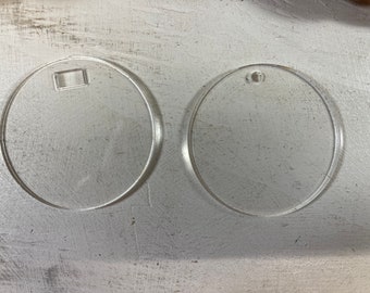 May include: Two clear, circular acrylic discs. The disc on the left features a small rectangular cutout near the top edge, while the disc on the right has a small circular hole positioned near the top edge for hanging or attachment purposes.