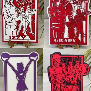 May include: Four layered wooden artworks depicting a marching band and a cheerleader. The pieces are red, white, black, and purple. Each piece has a name at the bottom: Izzy, Grady, Presley Peace, and Aiden. The artworks are displayed on wooden stands.