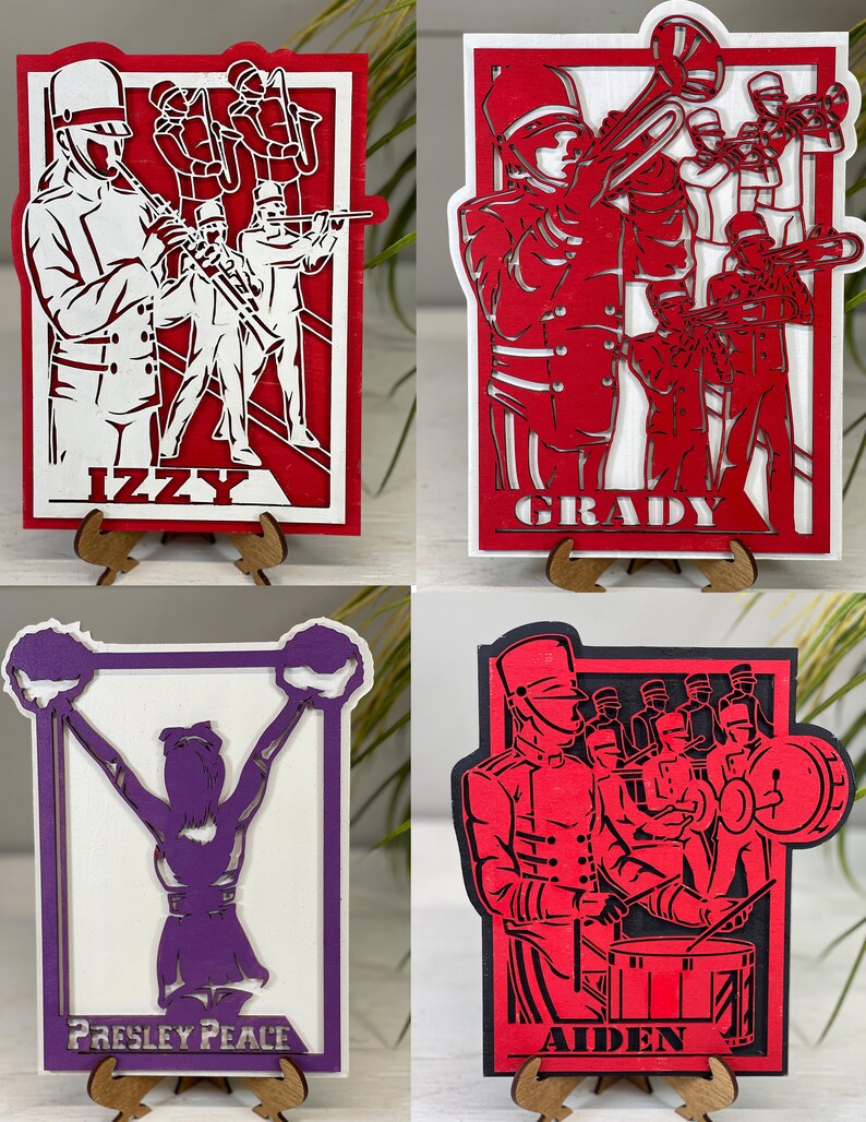 May include: Four layered wooden artworks depicting a marching band and a cheerleader. The pieces are red, white, black, and purple. Each piece has a name at the bottom: Izzy, Grady, Presley Peace, and Aiden. The artworks are displayed on wooden stands.
