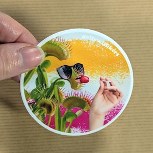 May include: A circular sticker featuring a collage of green Venus flytrap plants, a pair of black sunglasses, and a hand holding a red lollipop. The background transitions from yellow to pink with a textured effect and the text gollywalkers.