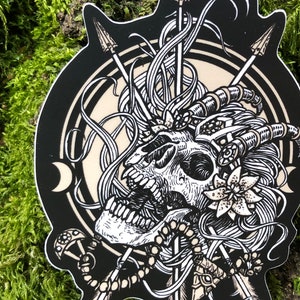 May include: A black and white sticker featuring a detailed skull with horns, arrows, and floral elements. The skull is the central focus, surrounded by intricate designs and a circular border. The sticker has a unique, die-cut shape.