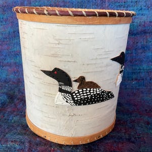 May include: A white birch bark trash can with a brown leather rim and a design of three black and white loons. The loons are painted on the side of the can.