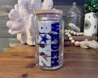 Puede incluir: Una lata de vidrio transparente con una tapa de madera. La lata tiene un diseño azul y blanco con el texto "WE DEM BOYS" y una estrella azul.