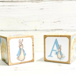 May include: Two wooden blocks with illustrations of Peter Rabbit. One block features Peter Rabbit wearing a blue jacket and the other block features the letter A with Peter Rabbit in a floral wreath.