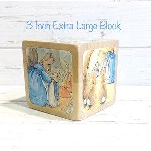May include: A 3 inch extra large wooden block with a Peter Rabbit illustration on each side. The block features Peter Rabbit in a blue dress, a brown rabbit with a blue jacket, and a group of brown rabbits.