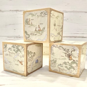 May include: Four wooden blocks with illustrations from Winnie the Pooh and the Hundred Acre Wood. Each block features a different character and location from the stories, including Tigger, Pooh, Kanga, and Roo. The blocks are a light brown color with white text and illustrations.