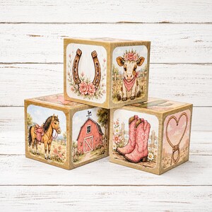 May include: Three wooden blocks featuring illustrations of a brown horse, a red barn, a horseshoe with pink flowers, a calf wearing a flower crown, pink cowboy boots, and a rope heart. The blocks have a light wood finish and decorative graphics.