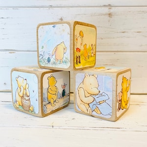 May include: Four wooden blocks with Winnie the Pooh illustrations. Each block features a different scene from the Winnie the Pooh stories. The blocks are stacked in a pyramid shape.