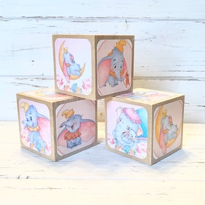 May include: Set of four wooden blocks featuring illustrations of Dumbo the elephant in various poses. The blocks are painted in soft pink, blue, and yellow tones with floral accents.