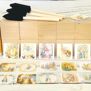 May include: A crafting set featuring a bottle of Mod Podge, paintbrushes, and wooden blocks. The set includes several small square cards with illustrations of rabbits in a storybook style. The Mod Podge bottle is 4 fl oz (118 ml).