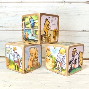 May include: Four wooden blocks with illustrations from Winnie the Pooh. The blocks feature Winnie the Pooh, Piglet, and Christopher Robin. The blocks are painted in a vintage style.