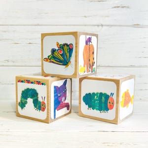 May include: Wooden toy blocks featuring colorful illustrations of a butterfly, a bear with a cat, a caterpillar, a cat, and a fish. The blocks are square with rounded corners and are made of light-colored wood.