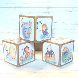 May include: Set of four wooden blocks with illustrated scenes from classic children's stories. Each block features a different character and scene, including a cow on the moon, a girl reading a book, a girl with sheep, and a man with a crown.
