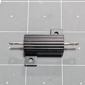以下が含まれることがあります： 黒いアルミニウム製ハウジングの電力抵抗器。円筒形の溝付きヒートシンク本体の両側に銀色の平らな金属端子が突き出ており、上下の縁には円形の穴が開いた取り付けタブが備わっています。