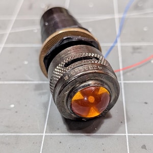 Puede incluir: Un componente de luz indicadora cilíndrico de metal con un acabado metálico oscuro, un agarre moleteado texturizado y una lente de vidrio de color ámbar prominente. El componente muestra el texto grabado 42B3-B e incluye dos cables finos.