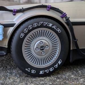 Puede incluir: Un modelo de coche plateado con neumáticos negros y llantas plateadas. Los neumáticos tienen letras blancas que dicen "Goodyear Eagle GT". El coche tiene cables negros con sujetadores morados.