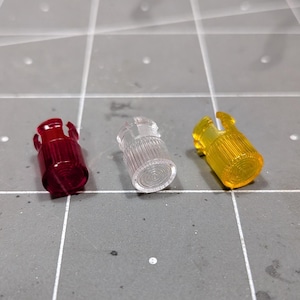 Peut inclure: Trois petits capuchons cylindriques en plastique translucide de couleur rouge, transparente et jaune. Chaque capuchon présente une texture extérieure nervurée et un bord supérieur cranté, conçu pour servir de couvercle ou de bouton.