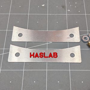 May include: Two rectangular silver metal plates with curved top edges and two circular holes each. One plate features the red text HASLAB. Included are two black metal bolts, two gold-colored threaded inserts, and two silver-colored metal hex nuts.