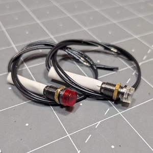May include: Two panel-mount LED indicator lights with black threaded metal housings and brass hex nuts. One light features a translucent red lens, while the other has a clear lens. Each unit includes a white cylindrical base and black wires.