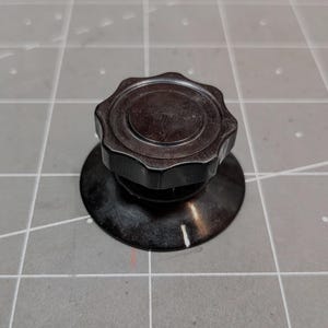 Puede incluir: Un mando de control circular de color marrón oscuro con un borde festoneado en forma de engranaje en la parte superior. La base es un disco plano y liso con una línea indicadora blanca marcada en el lateral, diseñado para su ajuste.