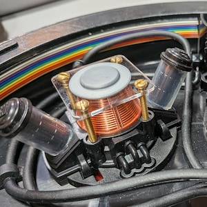 Puede incluir: Una vista interna de un dispositivo electrónico circular negro que presenta dos bobinas de alambre de cobre montadas sobre placas de acrílico transparente con tornillos de latón. El conjunto incluye varios componentes cilíndricos negros, cableado negro y un cable plano multicolor.
