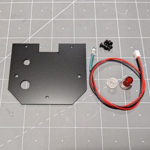 May include: A black metal plate with various holes, alongside small screws, a red LED, a clear plastic washer, and a red button with a silver base. Red and black wires are also present.