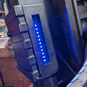 May include: A black electronic prop device featuring a vertical row of bright blue LED lights embedded in a rectangular housing. A multi-colored ribbon cable is secured by a black ring, and a red warning label with white text is visible.