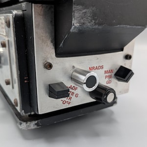 May include: A black and silver electronic control unit featuring a metallic faceplate with red text labels including NRADS, MAN, PRE, GD, ADF, TR G, TR, and OFF. The device includes several black and silver knobs and switches for manual operation.