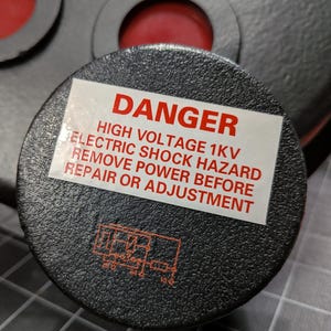 May include: A circular black textured component featuring a white warning label with red text that reads DANGER HIGH VOLTAGE 1KV ELECTRIC SHOCK HAZARD REMOVE POWER BEFORE REPAIR OR ADJUSTMENT and a small red schematic diagram printed below the label.