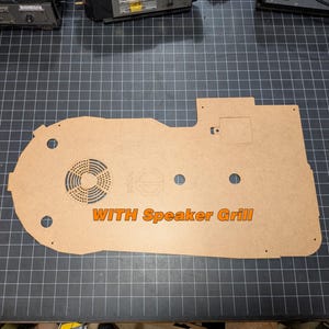 May include: A tan-colored acrylic panel with a speaker grill design and the text "WITH Speaker Grill" in orange. The panel has several circular cutouts and a rectangular cutout. The panel is on a black grid surface.