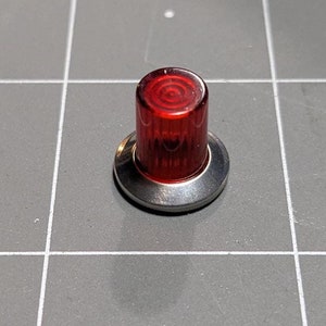 May include: A small, red, cylindrical light with a spiral design on top, mounted on a silver-colored base. The light is likely an indicator or a small warning light.