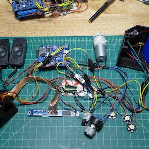 May include: A close-up of a workbench with various electronic components, including circuit boards, wires, speakers, a motor, and LEDs. The components are connected together with wires and are arranged on a green cutting mat.