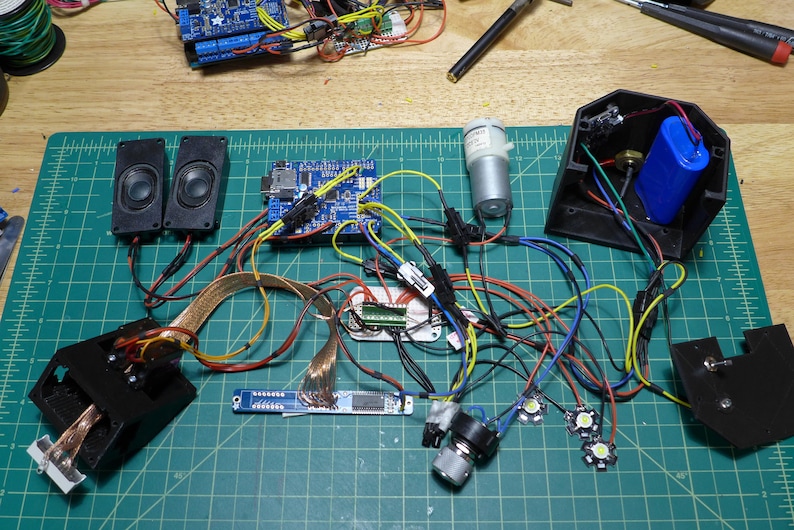 May include: A close-up of a workbench with various electronic components, including circuit boards, wires, speakers, a motor, and LEDs. The components are connected together with wires and are arranged on a green cutting mat.