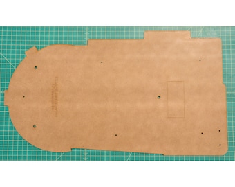 May include: A tan-colored, laser-cut wooden base with a rounded left side and a rectangular cutout. The base has several small holes and the text "CHARLESWORTH DYNAMICS" is printed on the left side. The base is on a green cutting mat.
