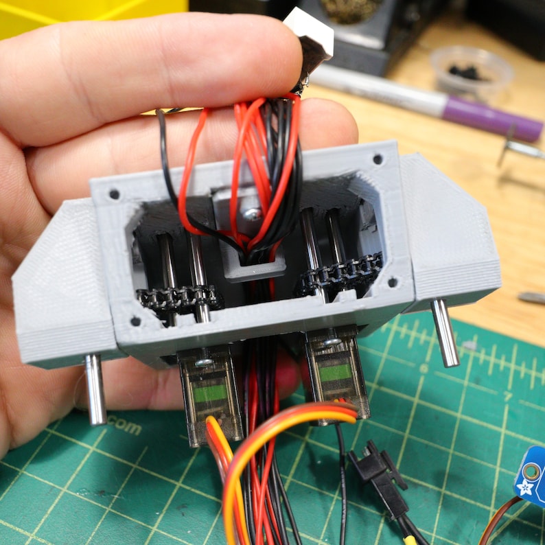 May include: A close-up of a 3D printed gray plastic device with two small motors and a chain drive system. The device is connected to a variety of wires, including red, black, and yellow wires.