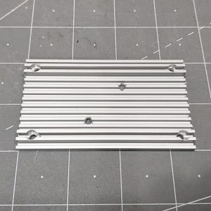 May include: A rectangular silver-coloured aluminium heat sink featuring a series of parallel longitudinal fins and five countersunk mounting holes distributed across the surface, designed for thermal management in electronic components.
