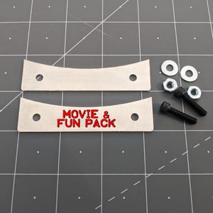 May include: Two curved silver metal plates with two circular holes each. One plate features the red text "MOVIE & FUN PACK". Included are two silver metal washers, two silver metal nuts, and two black metal bolts with hexagonal heads.